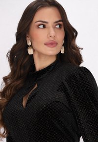 Bluză din catifea neagră cu guler înalt și decupaj în formă de cheie, având un model cu diamante format din pete colorate. Cercei texturați aurii.