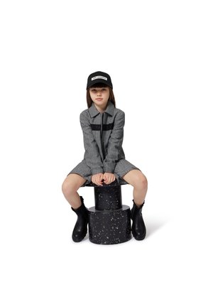 Robe tissée grise avec une fermeture éclair et un accent noir, associée à une casquette noire et des bottines noires, assise sur un piédestal cylindrique noir.
