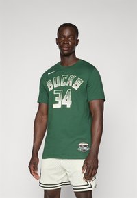 Žalias medvilnės marškinėliai su „BUCKS“ ir „34“ auksinės blizgesio spalvos, apvaliu kaklu, su Nike logotu ant kairiojo peties, derinami su baltais tinkliniais šortais.