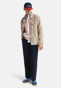 Beige Hemdjacke, rosa Pullover, hellblauses Hemd, marineblaue Hose und blaue Sneaker mit weißen Akzenten, dazu eine marineblaue und rote Kappe.