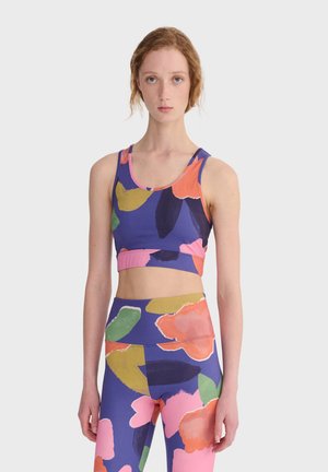 Mladá žena oblečená v zladenom farebnom kvetinovom športovom podprsenke a legínach s abstraktnými veľkými okvetnými lístkami na purpurovom pozadí.