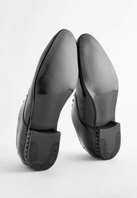 Chaussures noires brillantes avec des semelles en caoutchouc texturé, des orteils arrondis et des talons plats. Présentent une finition lisse et un dos structuré pour un soutien.