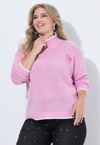 Roze gebreide trui met een witte kraag en manchetten, voorzien van een rode hartborduursel op de voorkant. Gecombineerd met donkerdenim jeans versierd met parels.
