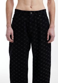 Pantalon en denim noir avec un motif de logo discret, coupe droite et style classique à cinq poches, garniture métallique sur la ceinture.