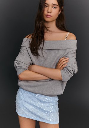 Junge Frau mit langem, dunklem Haar, die einen grauen, schulterfreien Pullover und einen hellblauen Minirock mit Pailletten trägt, steht mit verschränkten Armen.