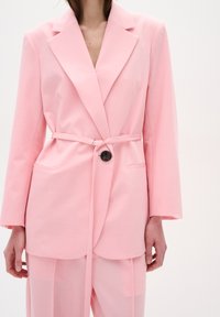 Rozā žakete, kas izgatavota no viegla auduma, ar jostu viduklī, dubulto pogu dizainu un lielām priekšējām kabatām ar melna pogu akcentu.
