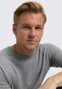 Blonde man met kort haar in een grijze trui, kijkt recht in de camera met een neutrale uitdrukking op een lichte achtergrond.