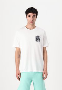 Camiseta branca com gola redonda e mangas curtas, apresentando um logótipo bordado a preto no lado esquerdo do peito. Tecido de algodão suave.
