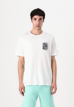 T-shirt blanc avec un col rond et des manches courtes, arborant un logo noir brodé sur la poitrine gauche. Tissu en coton lisse.