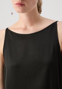 Top negru fără mâneci, realizat dintr-un material neted, cu un decolteu rotund, având o cusătură minimă și o croială lejeră. Cercei argintii în formă de inimă vizibili.