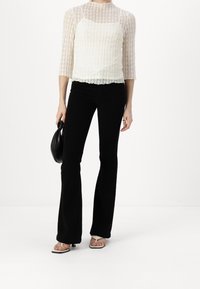 Blusa de encaje con mangas tres cuartos en color crema, combinada con jeans negros de campana y un bolso negro. Lleva sandalias de dedo de color claro.