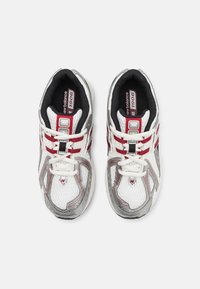 Baskets en mesh blanches avec des accents gris et rouges, présentant un design à lacets, un col rembourré et des superpositions texturées sur les côtés.