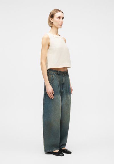 Femme debout, portant un haut court crème sans manches, un jean bleu délavé à jambes larges et des ballerines noires, sur un fond blanc uni.