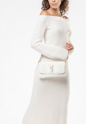 Frau in einem weißen schulterfreien Strickkleid, die eine weiße Umhängetasche mit goldenem V73-Logo vor weißem Hintergrund trägt.