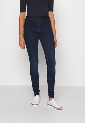 Jeans de mezclilla oscuros de tiro alto con una cinturilla flexible; forma ajustada, longitud hasta el tobillo, combinados con zapatillas blancas.