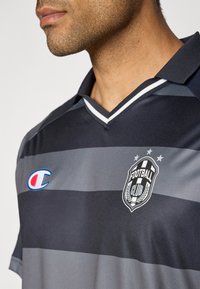 Grijze en donkergrijze gestreepte voetbaltrui met een V-hals, voorzien van een logo met laurieraccenten en een klein rood en blauw embleem.