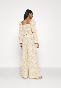 Macaquinho floral com mangas bufantes, decote à mostra dos ombros e design de pernas largas. Apresenta um tecido amarelo pastel com um padrão multicolorido.