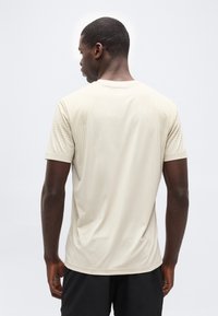 Άνδρας που φοράει μια απλή μπεζ κοντομάνικη μπλούζα και μαύρο σόρτς, στέκεται γυρισμένος με την πλάτη σε ένα απλό φωτεινό φόντο.