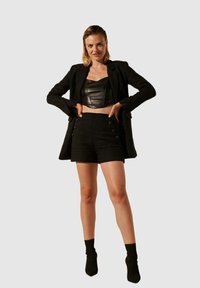 Blazer noir court porté sur un bustier en cuir noir et un short texturé taille haute avec des boutons décoratifs. Le mannequin porte des chaussettes noires aux chevilles.