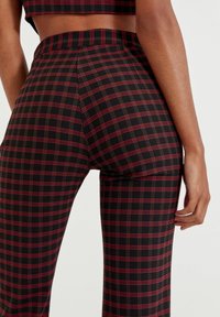 Zwarte high-waisted broek met een rood en groen ruitpatroon, voorzien van een getailleerd ontwerp en een gladde textuur, die de vorm en pasvorm benadrukt.