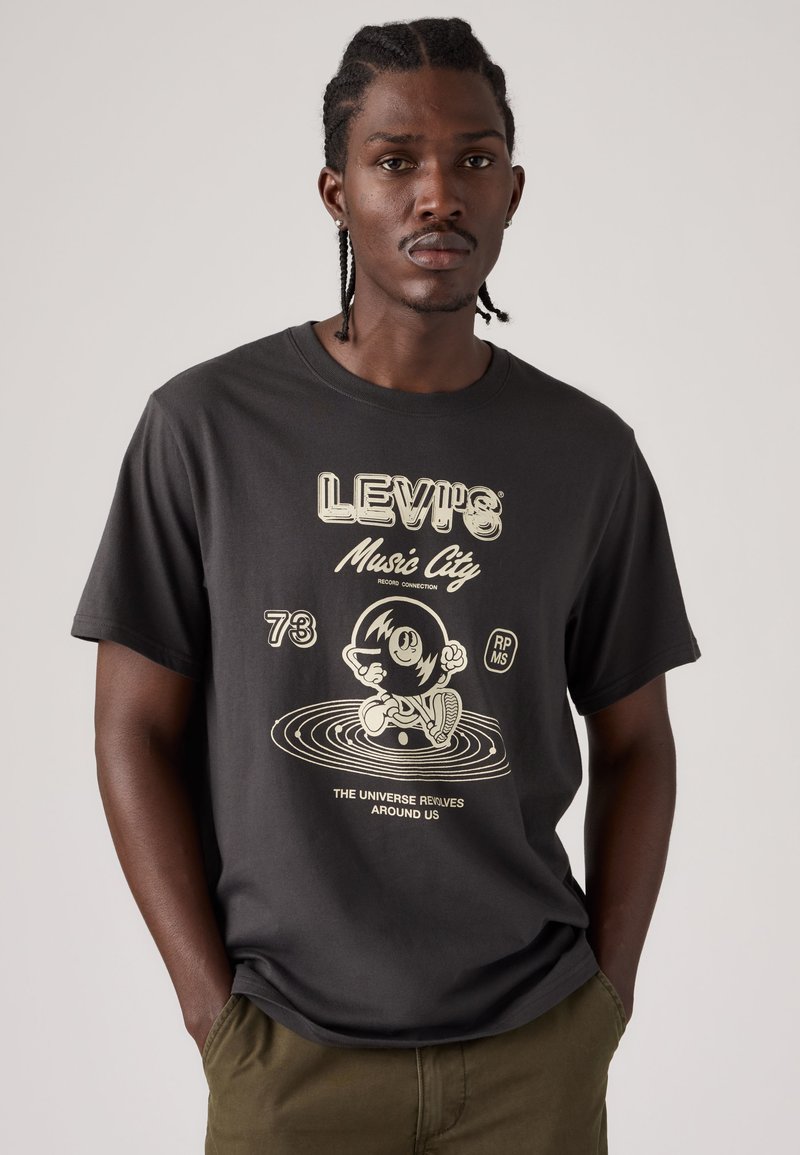 Hombre con cabello trenzado que lleva una camiseta negra de Levi’s con un disco de vinilo de dibujos animados y el texto "Music City" y "El universo gira a nuestro alrededor."