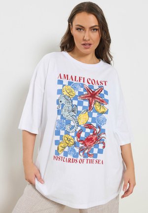 Frau trägt weißes übergroßes T-Shirt mit blau kariertem Druck, der Seepferdchen, Seesterne, Krabben, Zitronenscheiben und den Text "Amalfi Coast Postcards of the Sea" zeigt.