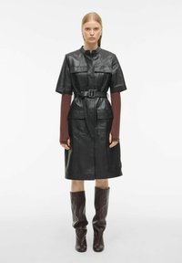 Juoda odinė suknelė su trumpomis rankovėmis, diržu talijoje ir priekinėmis kišenėmis. Ruda odinė batas iki kelių. Paprastas dizainas ir struktūruota forma.