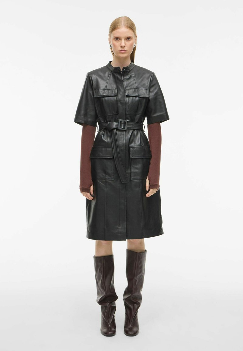 Juoda odinė suknelė su trumpomis rankovėmis, diržu talijoje ir priekinėmis kišenėmis. Ruda odinė batas iki kelių. Paprastas dizainas ir struktūruota forma.