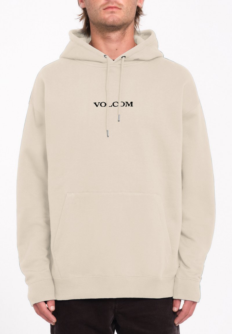Haut à capuche beige avec une poche kangourou à l'avant et un cordon de serrage, présentant un logo en texte noir "VOLCOM" sur la poitrine. Texture de tissu douce.