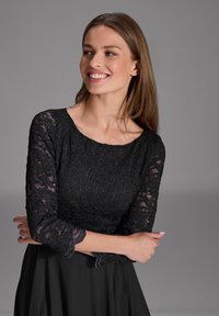 Robe en dentelle noire avec des manches trois-quarts, présente un corsage en dentelle ajusté et une jupe fluide en noir uni, texture lisse tout au long.