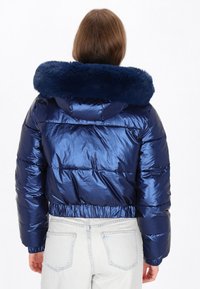 Námorně modrá péřová bunda se lesklou, prošívanou tkaninou a umělým kožešinovým límcem. Má přiléhavý pas a dlouhé rukávy.