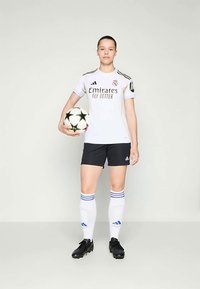 Balts futbola krekls ar melniem šortiem, uz kura ir melns "Emirates FLY BETTER" logo, apvienots ar baltiem zeķēm un melniem futbola apaviem. Tur rokās melnbalts futbola bumba.