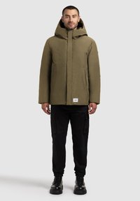 Man wearing olive green hooded winter coat, black cargo pants, and black lace-up boots standing against plain background.