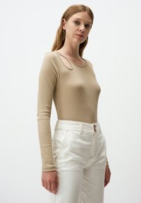 Beige langärmliger Body mit subtiler Aussparung an der Schulter, geripptem Muster, kombiniert mit hoch geschnittenen weißen Jeans mit zwei Knöpfen.