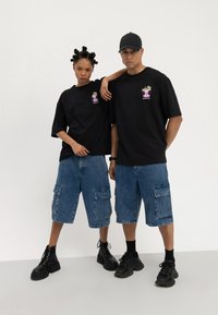 Camisetas negras de gran tamaño con un gráfico floral, combinadas con pantalones cortos cargo de mezclilla azul. Ambos llevan calzado negro para un look casual.