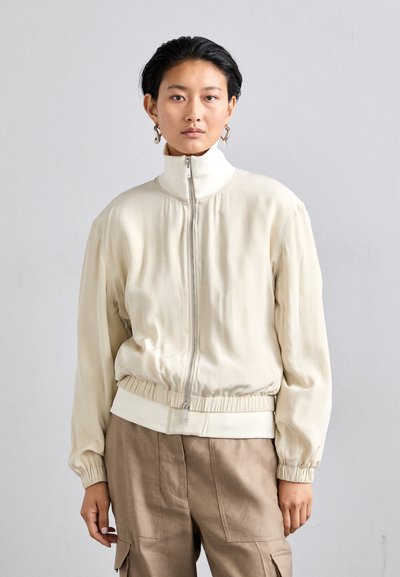 Veste bomber zippée en crème clair avec un col haut, des poignets élastiques et un ourlet froncé. Tissu lisse avec une coupe confortable et un design minimaliste.