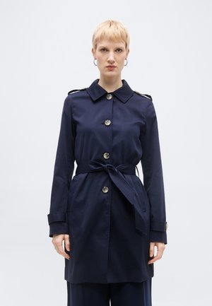 Kvinde med kort blondt hår iført en marineblå trenchcoat med bælte og store knapper samt øreringe i form af runde hoops, stående mod en ensfarvet baggrund.