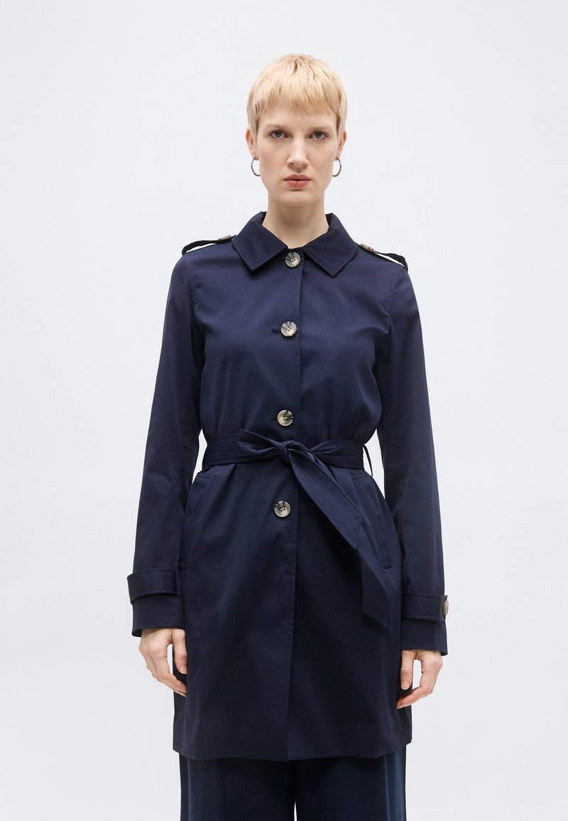 Γυναίκα με κοντά ξανθά μαλλιά, που φοράει μπλε ναυτικό παλτό με ζώνη και μεγάλα κουμπιά, συνοδευόμενο από σκουλαρίκια κρίκοι, στέκεται μπροστά σε απλό φόντο.