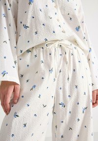 Witte pyjama set met een textielstructuur en blauw bloemenpatroon, knoopsluiting bovenstuk met lange mouwen en broek met elastische tailleband.