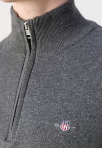 Gros plan d’un pull gris à fermeture éclair avec un col côtelé et un petit logo GANT brodé en rouge, blanc et bleu sur la poitrine.