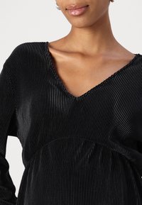 Musta, nauhomaista kangasta oleva mekko V-pääntiellä ja pitkillä hihoilla. Rakennemateriaalissa on pystysuoria raitoja ja se kerääntyy vyötärölle.