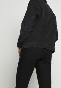 Persona vestida con una chaqueta de mezclilla negra ajustada y jeans negros, mostrada desde la mitad de la espalda hasta la mitad del muslo, contra un fondo claro liso.