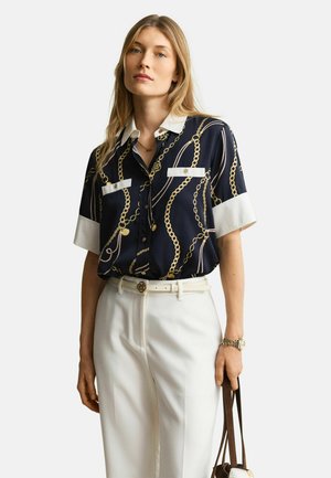 Vrouw die een marineblauw shirt met korte mouwen draagt met een gouden kettingprint, witte kraag en zakafwerking, witte broek met hoge taille en een bruine handtas vasthoudt.