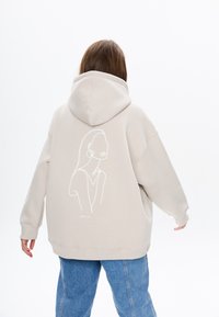 Beige oversized hoodie met een trekkoord capuchon. Heeft een witte lijntekening op de achterkant. Geribbelde manchetten en zoom. Gecombineerd met lichtblauwe jeans.