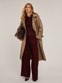 Beżowy trench coat na bordowym prążkowanym kardiganie i szerokich spodniach, trzymający ciemnobrązową skórzaną torbę. Gładkie tekstury, klasyczny fason.