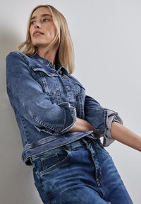 Ljusblå denimjacka med knäppning, två bröstfickor och uppvikta ärmar, kombinerad med mörkblå denimjeans.