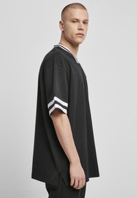 Černé oversized tričko s bílými a černými pruhovanými detaily na rukávech, s výstřihem do V a postranními rozparky na lemu. Hladká textura.