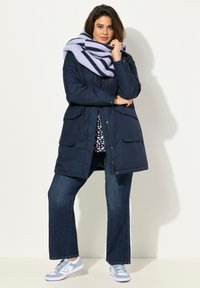 Marineblauer gepolsterter Mantel mit Fronttaschen, kombiniert mit dunkelblauen Schlagjeans und hellblauen Sneakers. Model trägt einen gestreiften lavendelfarbenen Schal.