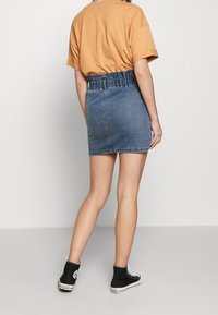 Jupe en denim mini avec ceinture élastique froncée, couleur bleu moyen, portée avec un t-shirt orange surdimensionné et des baskets noires montantes.