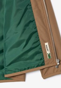 Veste marron avec doublure intérieure verte, partiellement dézippée, montrant une fermeture éclair argentée et une étiquette logo Lacoste avec emblème de crocodile vert.